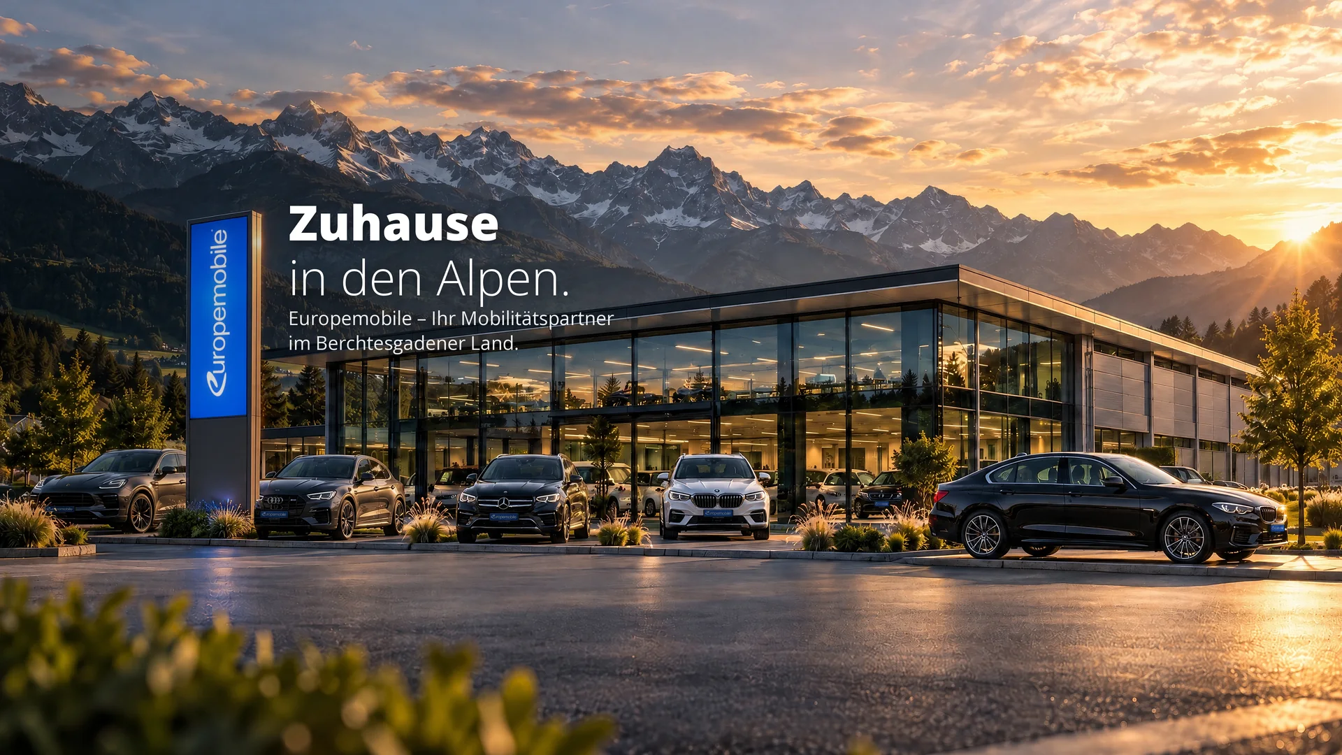 Zuhause in den Alpen: Europemobile-Autohaus mit Pylon und Fahrzeugen vor Bergpanorama bei Sonnenuntergang